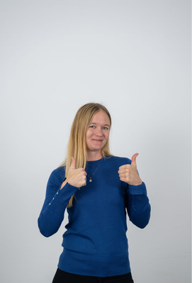 Junge Frau mit langem blondem Haar in blauem Pullover zeigt mit beiden Händen den Daumen nach oben und lächelt in die Kamera.