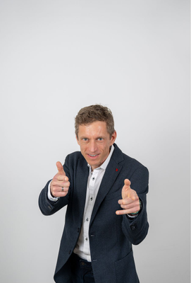 Mann in dunkelblauem Sakko und weißem Hemd, der lächelnd in die Kamera blickt und mit beiden Händen eine Fingerpistolen-Geste macht.