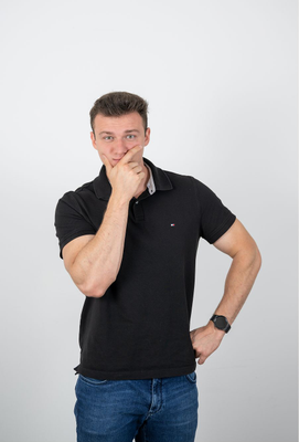 Junger Mann mit nachdenklichem Gesichtsausdruck, trägt ein schwarzes Poloshirt und blaue Jeans, stützt das Kinn mit einer Hand und blickt leicht zur Seite.