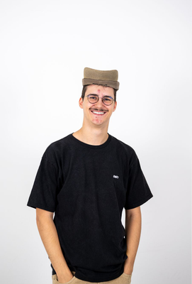 Lächelnder junger Mann mit Brille, Schnurrbart und schwarzem T-Shirt steht vor weißem Hintergrund, trägt eine umgedrehte Kappe auf dem Kopf.