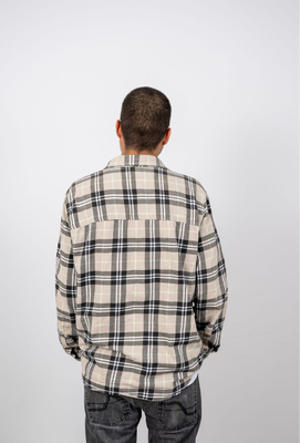 Mann mit kurz rasierten Haaren von hinten, trägt ein beige-grau kariertes Hemd und dunkle Jeans vor neutralem Hintergrund.