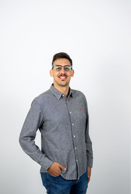 Junger Mann mit Brille und kurzem dunklem Haar lächelt leicht, trägt ein graues Hemd und Jeans, steht entspannt mit einer Hand in der Hosentasche vor hellem Hintergrund.